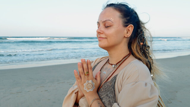 Woman standing with hands in namaste gesture and eyes closed on a sandy beach, focusing on spiritual meditation and calm mindfulness with the ocean waves in the background