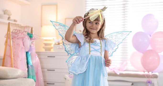 Cute little girl in fairy costume with wings, headband and magic wand sprinkling glitter at home