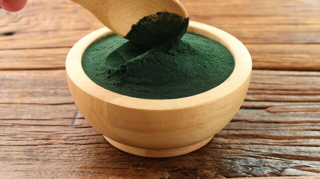 Woman taking spirulina powder with spoon from bowl on wooden table, closeup
