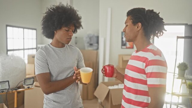 Man holds yellow mug and talks with friend near stacked moving boxes and packed furniture in a building; moving day camaraderie.