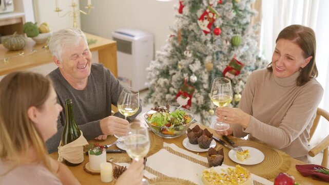Senior parents and adult daughter enjoy Christmas dinner together at home. Happy family sharing festive holiday meal with roast chicken, salad and wine around dining table during seasonal celebration.