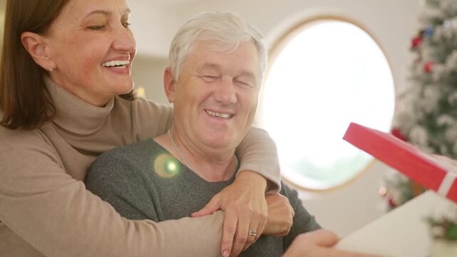 Family celebrating Christmas at home. Senior wife gives gift to husband while relaxing together in living room. Happy Caucasian older couple sharing love, surprise and festive holiday tradition.