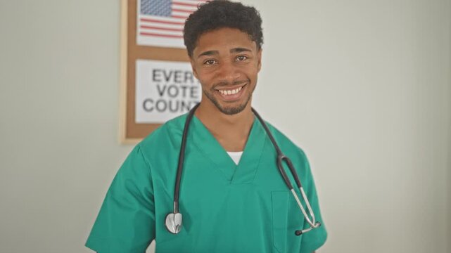 Man doctor in green scrubs with stethoscope points finger to camera in building by a vote poster and american flag; civic pride.