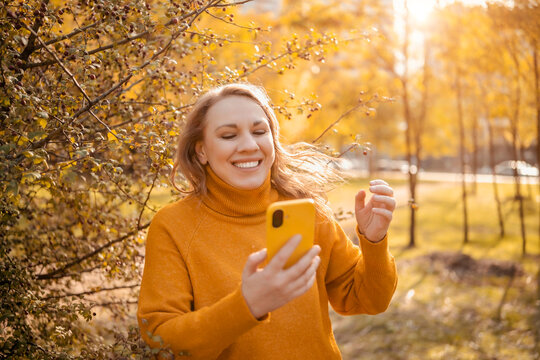 Joyful woman in autumn park with phone &mdash; digital communication concept. Happy adult messaging friends at fall outdoor leisure. Cheerful person laughing near sunny golden scenic bushes.