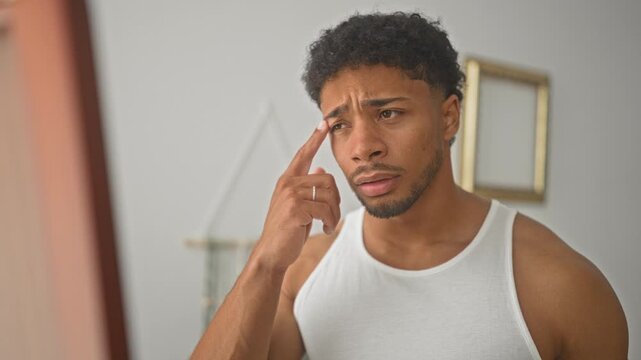 Man touching cheek and pointing at blemish while examining skin in bedroom mirror wearing white tank top and ring; skin care concern.