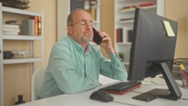 Man holds smartphone to ear while seated at desk with computer monitor, keyboard and paperwork in office building; quiet concentration.
