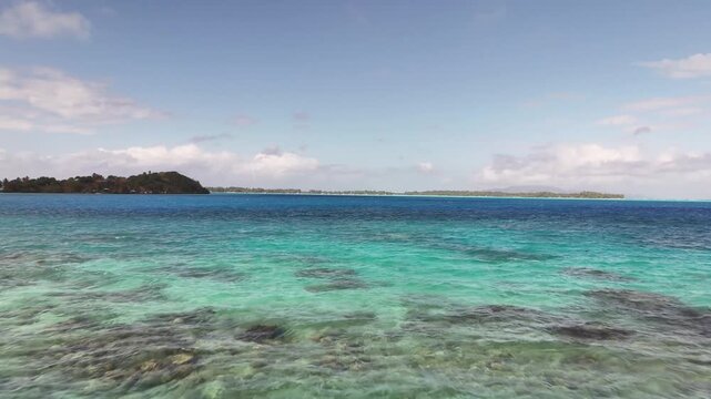 Aerial drone shot moving forward over a multi-shaded turquoise lagoon reef system, revealing a green coastal islet and distant flat sand motus of Bora Bora