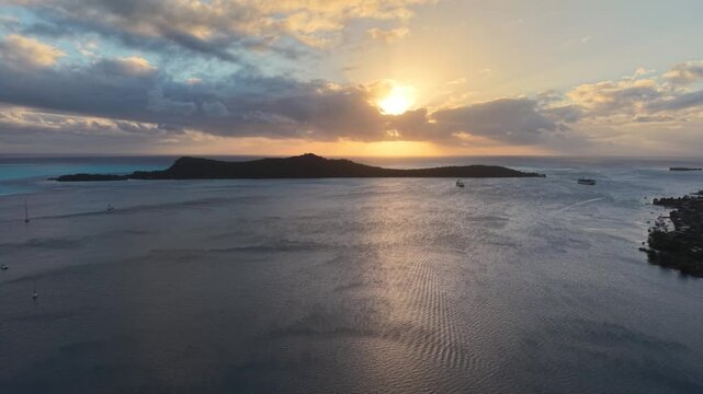 Aerial drone shot moving forward over calm lagoon waters at sunset, showcasing anchored boats and the silhouette of Motu Toopua island in Bora Bora