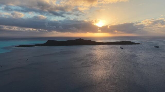 High-altitude aerial drone orbit shot tracking over a calm lagoon at sunset, showcasing anchored boats and the silhouette of Motu Toopua island in Bora Bora