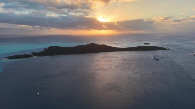 Aerial drone shot moving forward and rising over calm lagoon waters at sunset, showcasing an anchored ship and the silhouette of Motu Toopua in Bora Bora