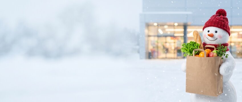 Minimalist winter panoramic banner: Cute smiling snowman holding a paper grocery bag with healthy fresh food. Blurred modern supermarket or clinic building background with falling snow. Perfect templa
