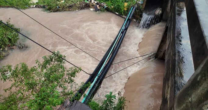 Slow motion of a turbulent brown river after heavy rain with blue and black  pipes and electrical wires crossing it next to a small bridge.