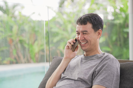 Man, smartphone, talking, smiling, outdoor, pool, relaxation - Happy middle-aged Asian man enjoying a casual conversation on his mobile phone while sitting on a terrace by the water.
