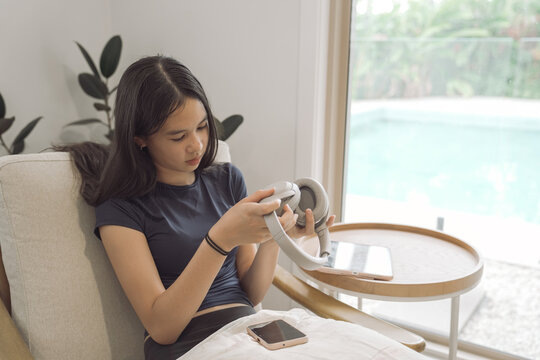 teenage girl, headphones, wireless headset, asian ethnicity, technology, lifestyle, Gen Z sitting in a cozy armchair indoors, examining her audio device for music and relaxation.
