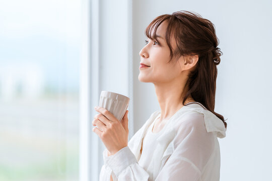 大きな窓のある明るいリビングでくつろぐ40代50代女性。自然光が差し込む快適なマイホーム
