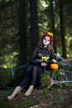 Young barefoot woman in the forest on a stump with a halloween pumpkin and yellow roses in hands.