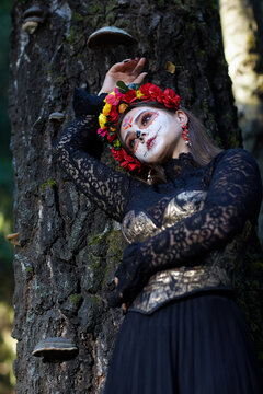 Halloween beauty portrait of female near tree trunk. Beautiful woman with Santa Muerte makeup.
