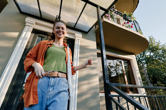 Smiling young woman in casual clothes standing on house porch near front door. Low angle view on sunny day. Concept of home, lifestyle, and happiness.