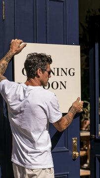 Opening Soon Sign: A man with tattoos hangs an Opening Soon sign on a blue door, signaling the imminent launch of a new business or establishment.