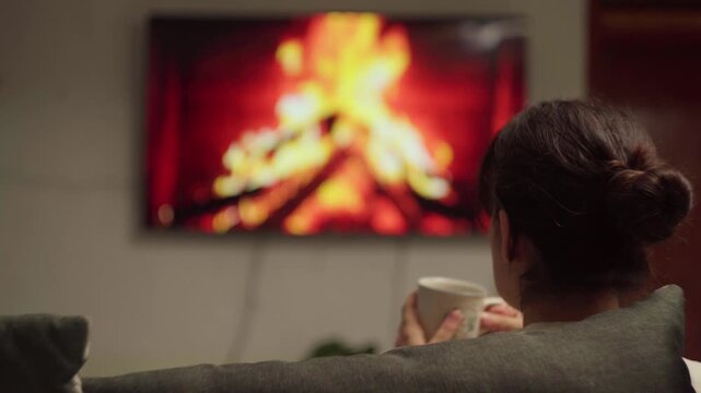 Woman drinking tea while watching fireplace video on smart TV in cozy living room at night