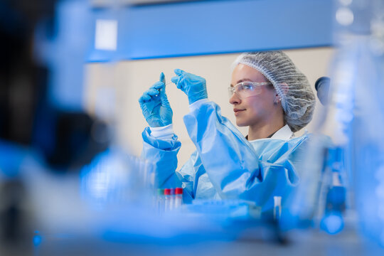 Focused female scientist in blue protective gown preparing a glass slide for microscopic analysis in sterile lab for clinical pathology, biotechnology research, and medical care.