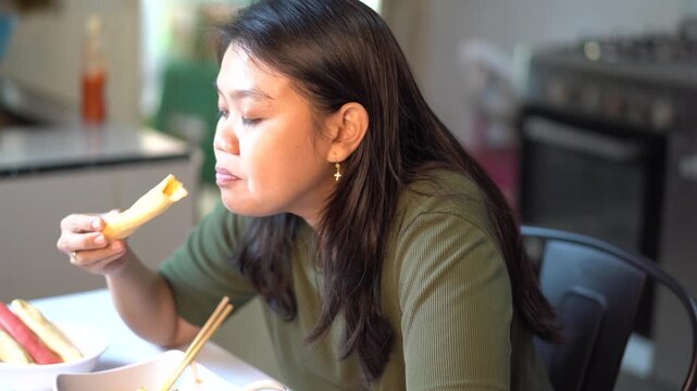 Asian woman eating spring roll with chopsticks in kitchen dining room