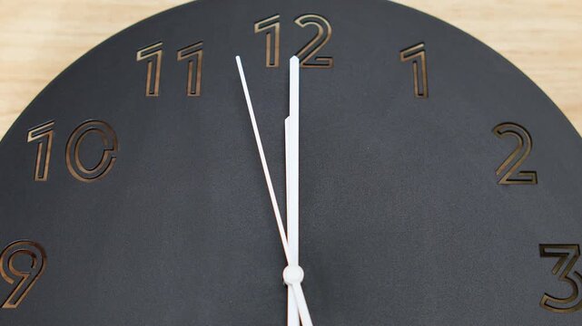 4K Close up shot of modern black wall clock face with engraved numbers and white hands pointing near twelve o'clock, time management and deadline concepts.