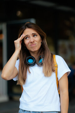 Confused Young Woman With Headphones Touching Forehead Being Very Forgetful. Unsure female student winces while trying to remember something important outdoors