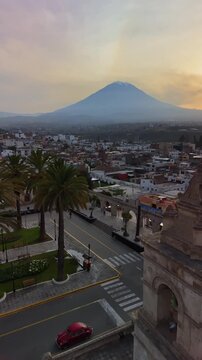 Vertical aerial drone view of the Misti volcano rising over the city, seen past the white sillar arches and palm trees of the Yanahuara viewpoint, Arequipa, Peru