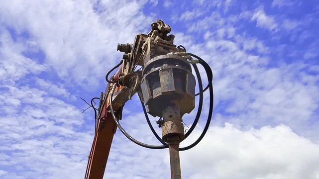 Hydraulic earth auger attachment on excavator arm for foundation drilling at construction site against blue sky.