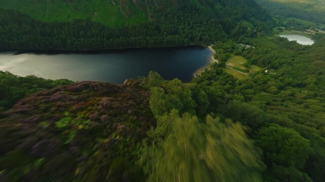 FPV camera glides along a dark mountain lake past forested hills and a sandy Glendalough beach with a few people on the shore.