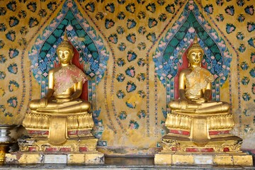 Obraz premium Two seated gold Buddha statues inside a cloister at Wat Arun, the Temple of Dawn in Bangkok, Thailand clothed in traditional Thai silk garments with ancient painted backdrop