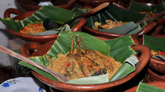 Traditional spicy eggplant curry inside an authentic clay pot lined with fresh green banana leaf using rustic wooden ladle spoon for indonesian culinary menu.