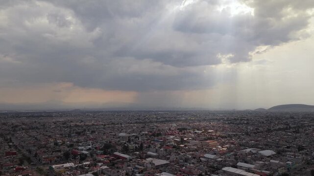 Aerial drone view of Ecatepec, Mexico, with urban buildings under cloudy skies, sun rays breaking through clouds and rain falling over the city in the distance