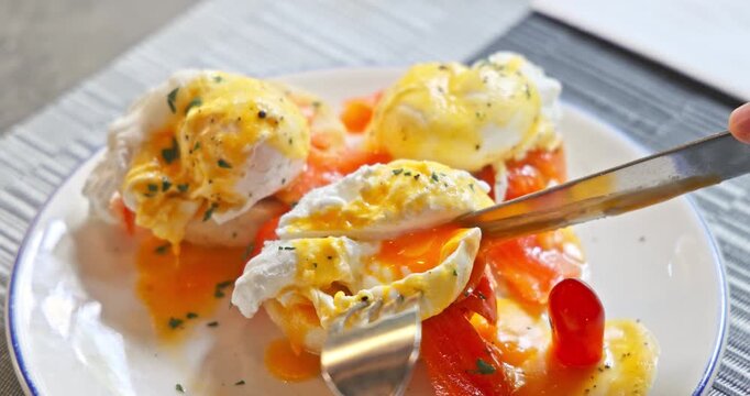 Delicious eggs benedict with smoked salmon and hollandaise sauce on a plate. Person cutting into a runny poached egg with a knife and fork. Gourmet breakfast meal in a restaurant setting.