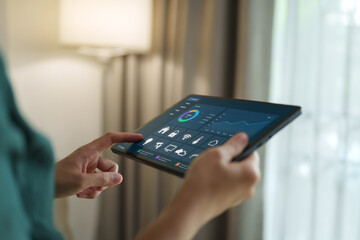 Woman using a digital tablet to control a smart home system, displaying a digital dashboard for energy monitoring, connected devices, automation, lighting management and efficient household living. © eggeeggjiew