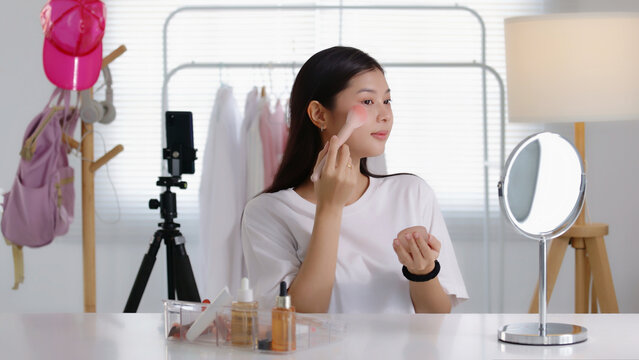 Young Asian beauty influencer applying blush with a powder brush while recording a makeup tutorial on smartphone in her room, perfect for cosmetics, blogging, and social media content.