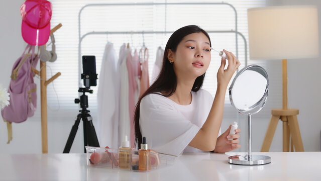Close-up of a young Asian woman applying eyeshadow with a small makeup brush in front of a mirror, ideal for cosmetics, beauty tutorials, and skincare content.