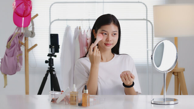 Young Asian beauty influencer applying blush with a powder brush while recording a makeup tutorial on smartphone in her room, perfect for cosmetics, blogging, and social media content.