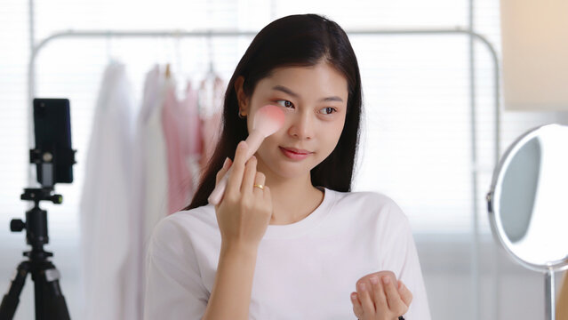 Young Asian beauty influencer applying blush with a powder brush while recording a makeup tutorial on smartphone in her room, perfect for cosmetics, blogging, and social media content.