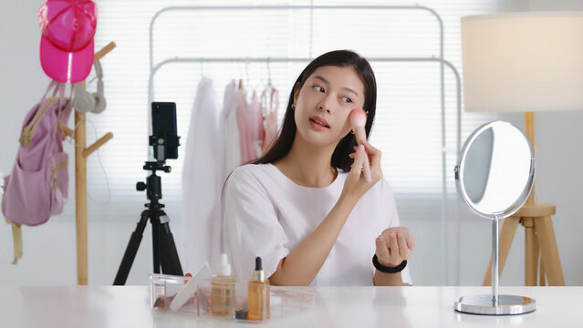 Young Asian beauty influencer applying blush with a powder brush while recording a makeup tutorial on smartphone in her room, perfect for cosmetics, blogging, and social media content.