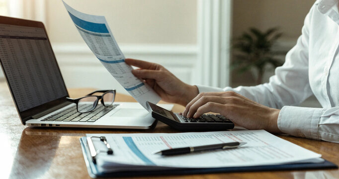 Accountant calculating financial report data with laptop and papers on wooden desk. Professional worker reviewing paper invoice with calculator and laptop computer
