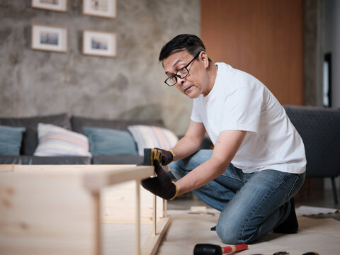 Asian mature man determinedly assembling wooden furniture with DIY tools, checking work precision with spirit&nbsp;level, home improvement, craft installation, hobby carpentry workshop, active lifestyle.