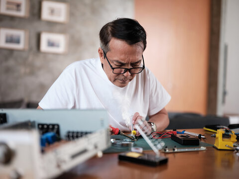 Mature Asian man repairs home electrical appliance, disassembling and soldering circuit board, focusing on precision detail, DIY fix tools, electronics component workshop hobby, expert handyman.