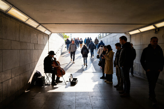Street musician playing cello in pedestrian tunnel while people listen, natural light from both ends