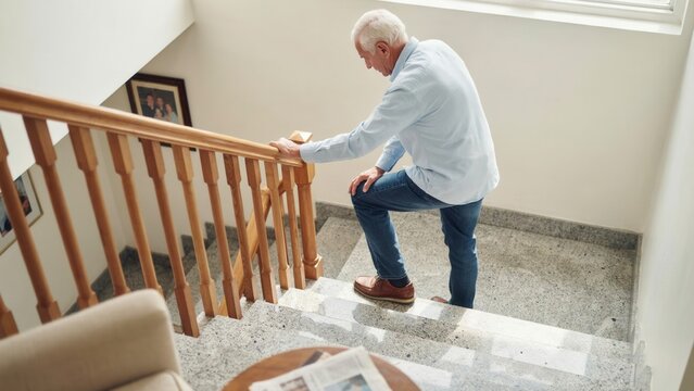 Elderly man walking carefully up indoor staircase using wooden handrail for support with physically active senior life wooden stairs elderly person balance support home life mobility aid
