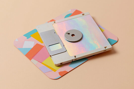 Pastel-colored floppy disk rests on a geometric mat. Soft pink, yellow, and blue shapes frame the nostalgic tech. Holographic finish catches the light gently. Slight wear suggests age and use