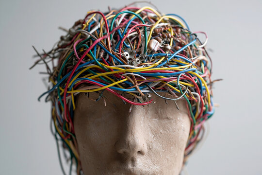 Tangled wires coil around a human head sculpture. Colorful cords drape over the face, forming a chaotic crown. The statue appears calm, eyes closed, expression serene
