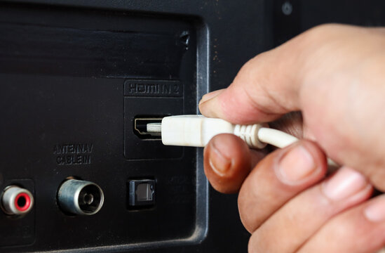 Close-up of a hand plugging a white HDMI cable into an HDMI port on the back of a television.