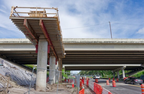 Bridge construction site on a highway overpass featuring scaffolding, concrete pillars, orange traffic barriers and cones, urban infrastructure work and road maintenance under a blue sky with clouds.
